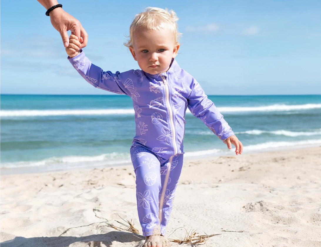 A toddler walking on a sandy beach holding an adult's hand, wearing a light purple full-body swimsuit with a white floral pattern and white zipper.