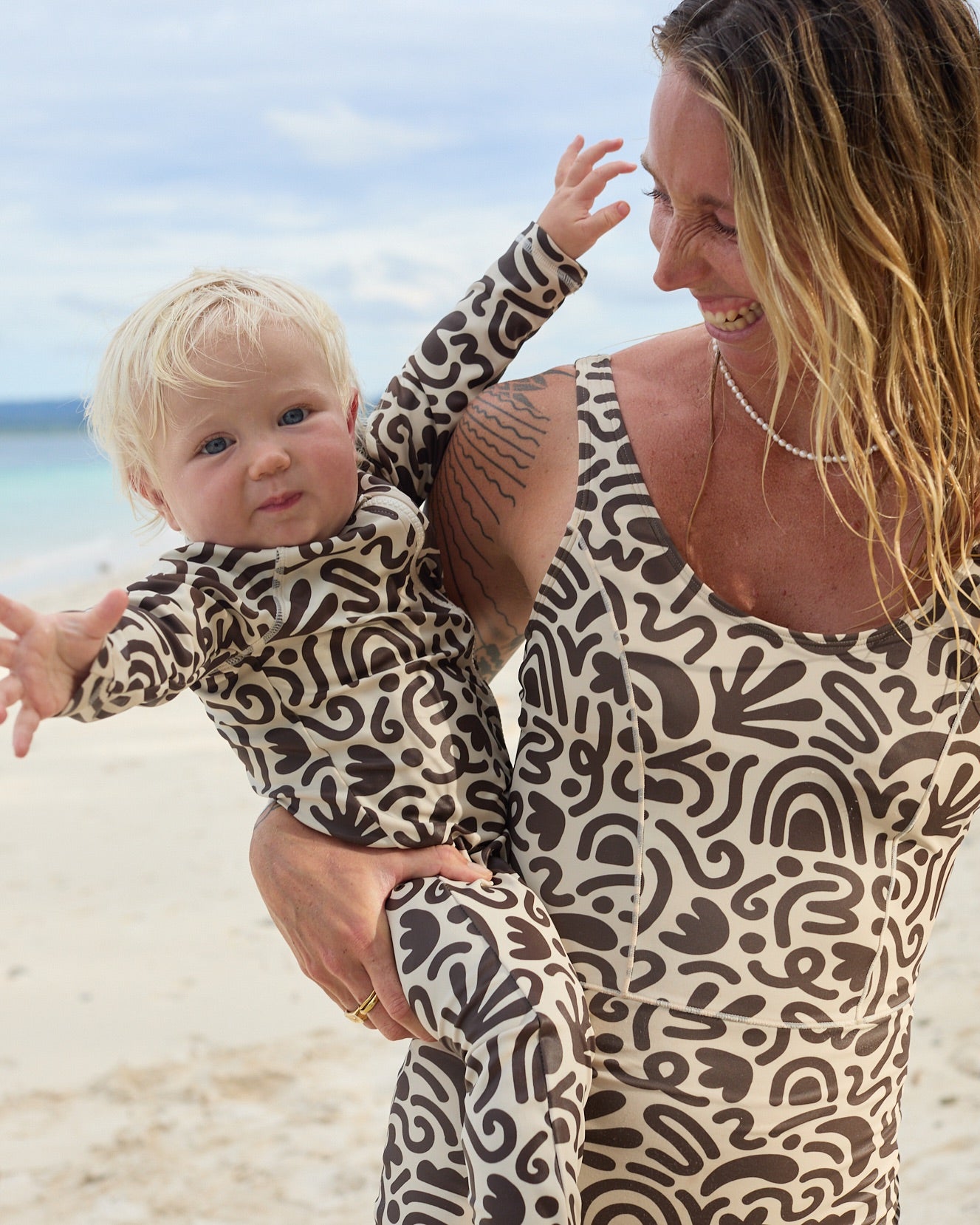 A woman and a toddler wearing matching long-sleeve patterned swim suits pose together on a sandy beach. The suits feature an abstract organic pattern in dark brown on a warm beige background.