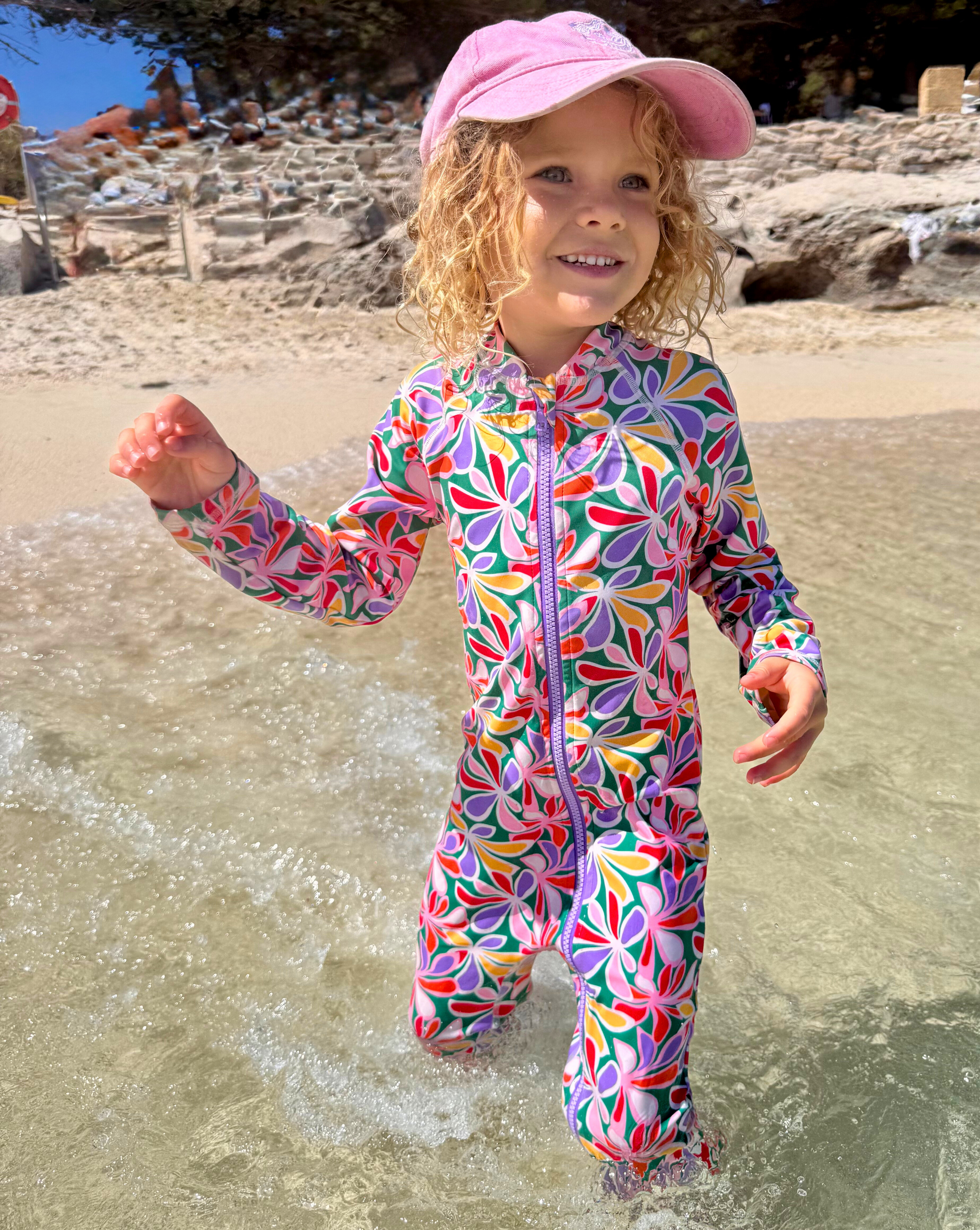 Child in a colorful swimsuit and pink cap standing in shallow water at the beach.